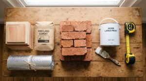 Flat lay of construction materials including bricks, tiles, paint can, cement bag, insulation roll and tape measure